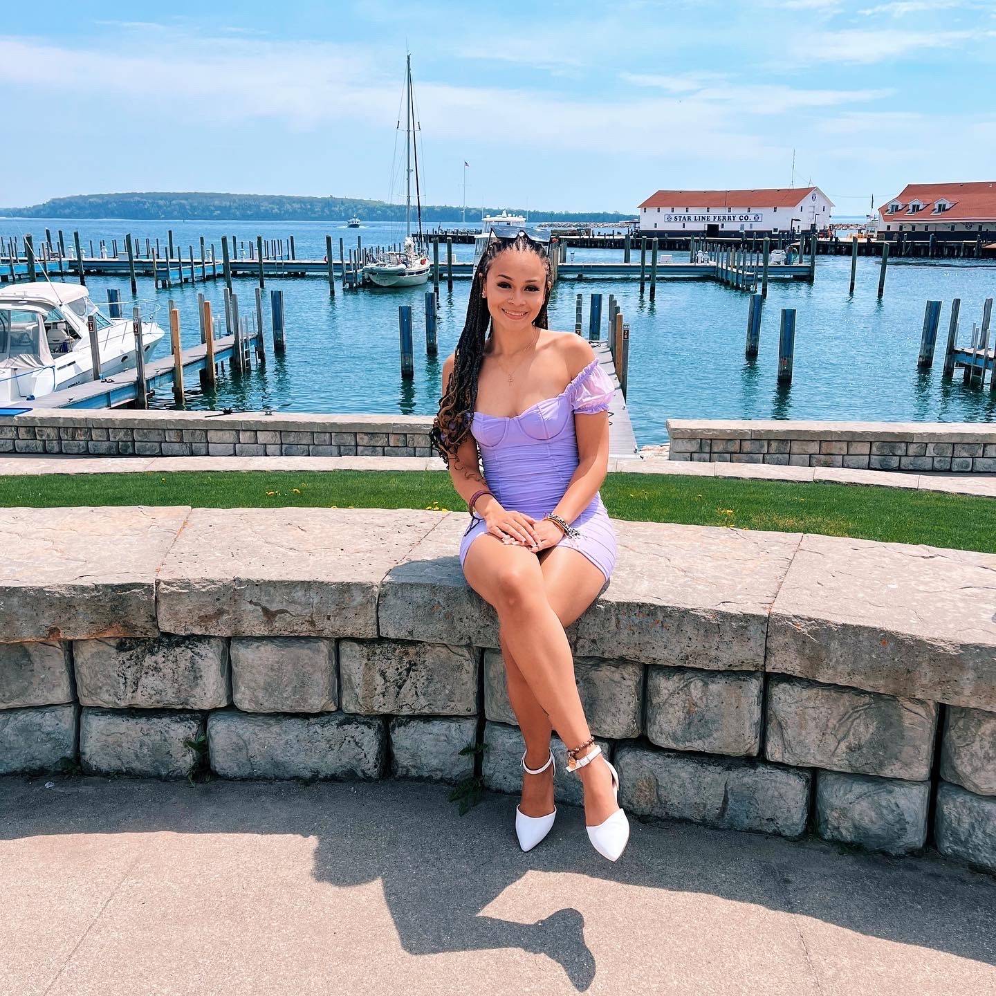 Girl with black hair and purple dress sitting on a stone wall with a lake and harbor in the background